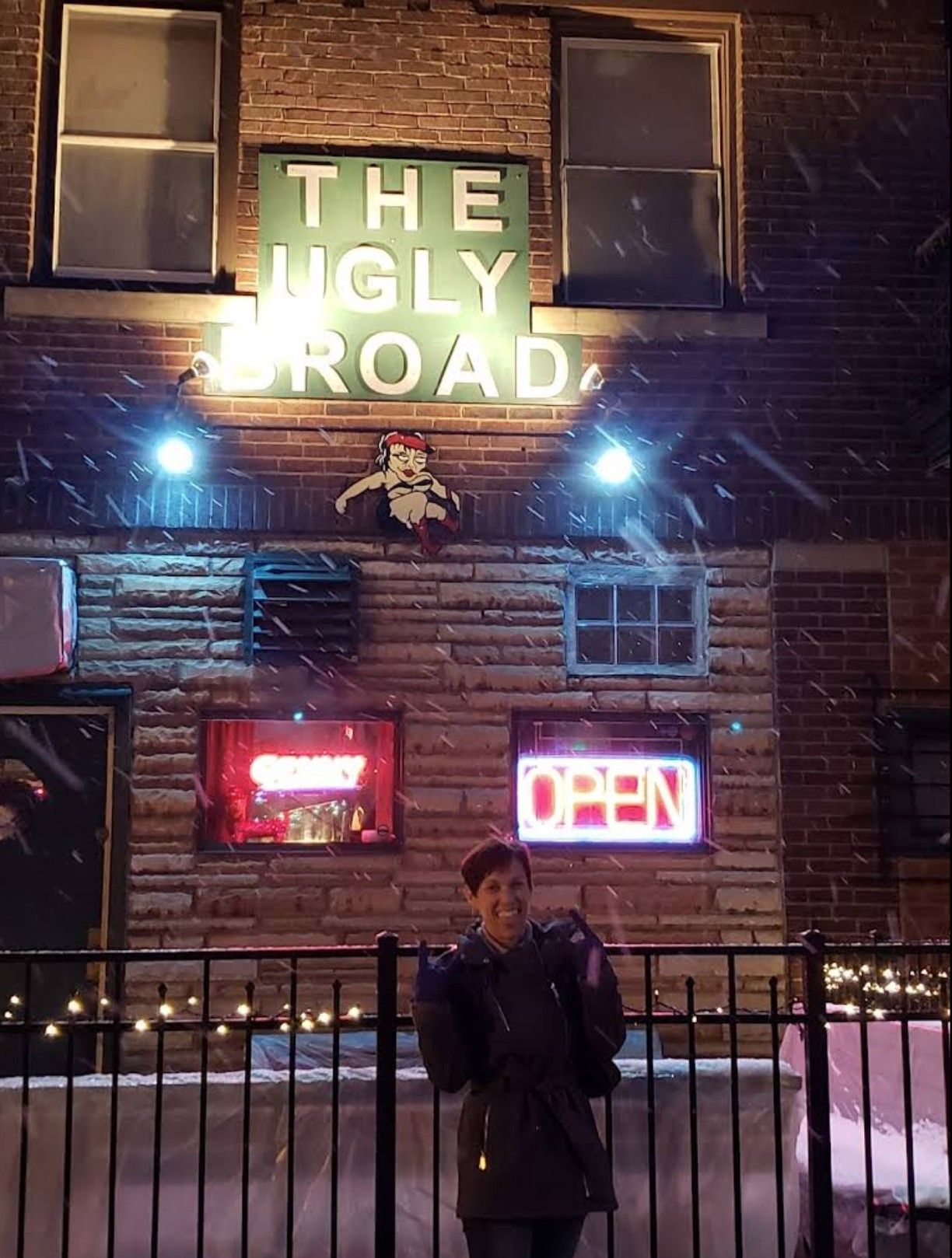 Exterior photo of a woman in front of The Ugly Broad sign while it snows.